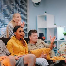 Happy viewers watching sports on TV and spilling popcorn when team scores. They are extremely happy, screaming, jumping and raising clenched fists.