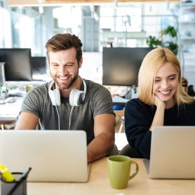 Photo of smiling young colleagues sitting in office coworking using laptop computers. Looking aside.
